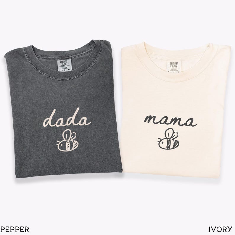 May include: Two folded t-shirts, one in pepper and one in ivory. The pepper shirt features the word "dada" and a bee illustration. The ivory shirt displays the word "mama" and a bee illustration. Both shirts are made of a soft fabric.