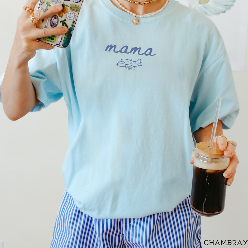 以下が含まれることがあります： ライトブルーのTシャツで、ダークブルーの筆記体で「mama」という文字と、その下に小さなダークブルーの飛行機のグラフィックが描かれています。青と白の縦縞模様のショートパンツと組み合わされており、シンプルでカジュアルなデザインの服装です。