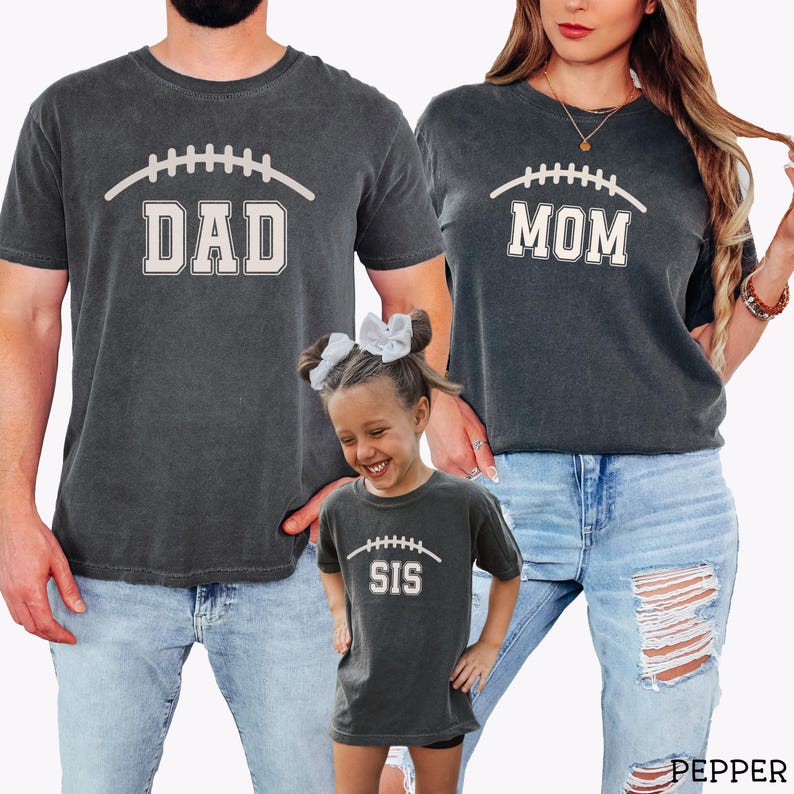 May include: Three charcoal gray t-shirts featuring a white graphic of a football lace design above the words DAD, MOM, and SIS in a collegiate block font. The shirts have a vintage-washed appearance and are worn with light blue denim jeans.