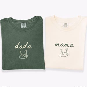 May include: Two t-shirts, one in moss green and one in ivory, with the words "dada" and "mama" embroidered on them. Each shirt features a small golf club and ball graphic.