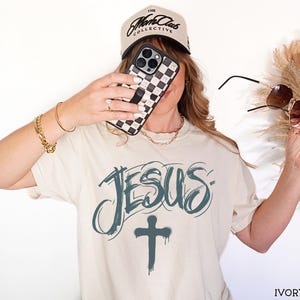 May include: Ivory-colored t-shirt with the word "JESUS" in teal script and a teal cross below. A black and white checkered phone case is held up. A beige baseball cap with black text is worn.