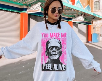 May include: A light gray crewneck sweatshirt featuring a graphic print of Frankenstein's monster centered on the chest. The design includes a bright pink rectangular background with the text YOU MAKE ME FEEL ALIVE in bold black and pink lettering.