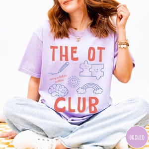 Peut inclure: Un t-shirt violet clair avec le texte rouge THE OT CLUB et la phrase every milestone matters. Le motif comprend des illustrations au trait bleu repr&eacute;sentant un crayon, deux pi&egrave;ces de puzzle, un cerveau, un soleil souriant et un arc-en-ciel.