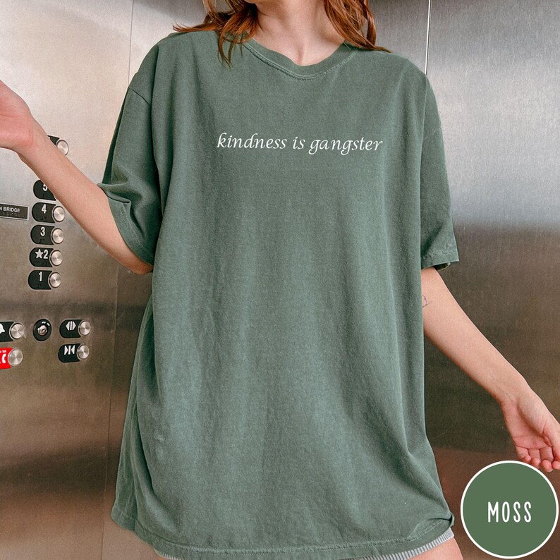 May include: A moss green t-shirt with the phrase "kindness is gangster" printed in white. The shirt has a relaxed fit and short sleeves. The image includes a color swatch labeled "MOSS".