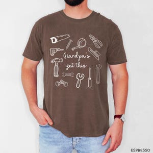 Peut inclure: T-shirt marron avec un motif blanc d'outils de bricolage, dont une scie, un marteau et une cl&eacute;. Le texte "Grandpa's got this" est imprim&eacute; au-dessus des outils. Le t-shirt est de style d&eacute;contract&eacute;.