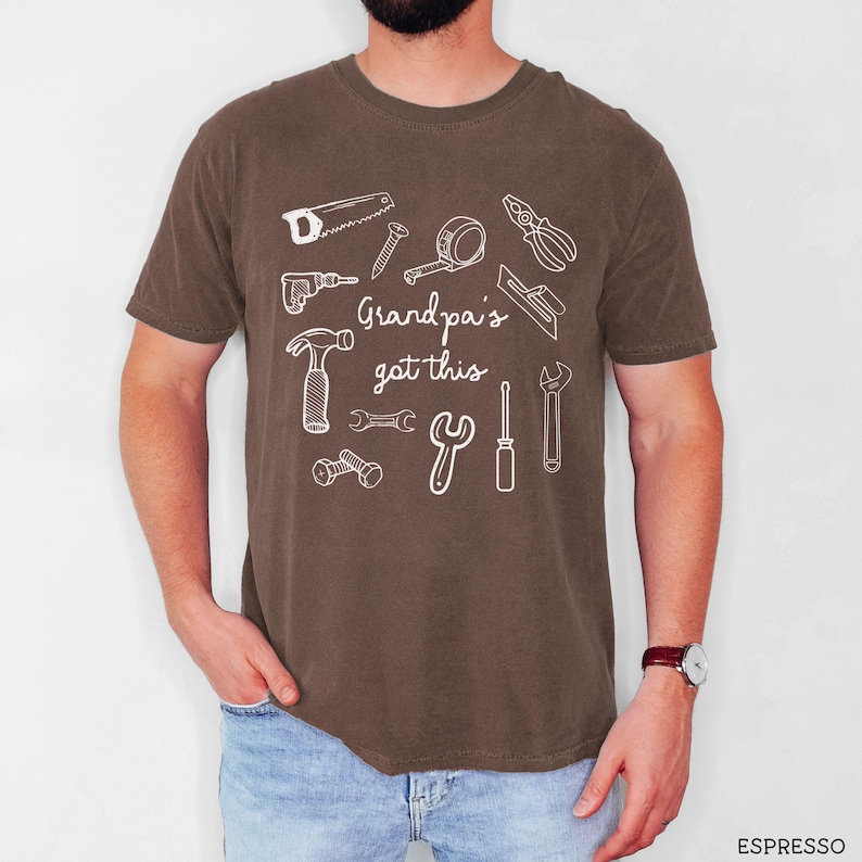 Peut inclure: T-shirt marron avec un motif blanc d'outils de bricolage, dont une scie, un marteau et une cl&eacute;. Le texte "Grandpa's got this" est imprim&eacute; au-dessus des outils. Le t-shirt est de style d&eacute;contract&eacute;.