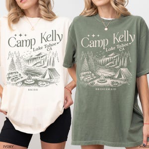 May include: Two short-sleeve crew neck t-shirts, one ivory and one moss green, featuring a dark green graphic print of a campsite with a tent, campfire, and forest. The text reads Camp Kelly Lake Tahoe CA, with BRIDE on the ivory shirt and BRIDESMAID on the moss green shirt.