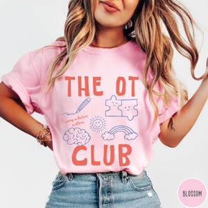 Peut inclure: Un t-shirt rose clair avec le texte THE OT CLUB en orange corail. Sous le texte se trouvent des illustrations au trait bleu repr&eacute;sentant un crayon, deux pi&egrave;ces de puzzle, un soleil, un cerveau, un arc-en-ciel et le texte every milestone matters.