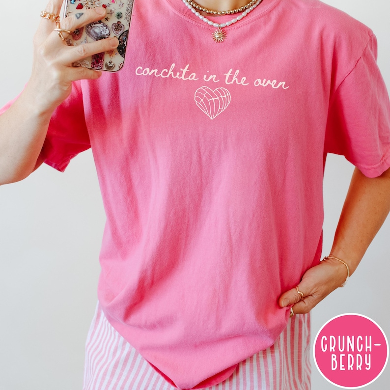 May include: A bright pink t-shirt with the phrase "conchita in the oven" in white script above a white heart graphic. The shirt is made of a soft material and is part of a clothing collection.
