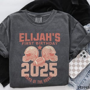Puede incluir: Una camiseta de manga corta gris carb&oacute;n con un gr&aacute;fico rojo desgastado que dice ELIJAH'S FIRST BIRTHDAY 2025 MAMA OF THE ROOKIE. El dise&ntilde;o incluye dos cascos de f&uacute;tbol americano y un bal&oacute;n de f&uacute;tbol americano centrados entre el texto y una estrella.