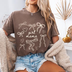 May include: A brown t-shirt with a white graphic design of bows and hearts. The text "girl mama" is centered on the shirt.