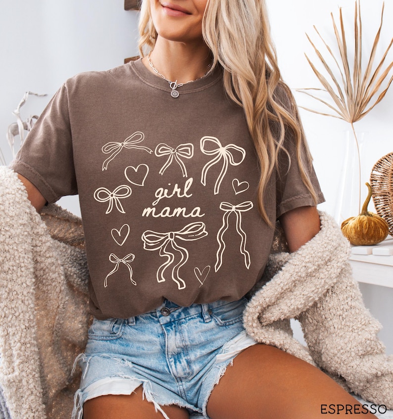 May include: A brown t-shirt with a white graphic design of bows and hearts. The text "girl mama" is centered on the shirt.
