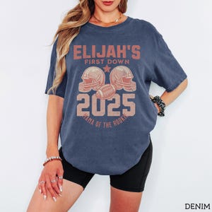 Puede incluir: Una camiseta azul marino de manga corta con un estampado gr&aacute;fico desgastado en rojo descolorido. El texto dice ELIJAH'S FIRST DOWN 2025 MAMA OF THE ROOKIE, acompa&ntilde;ado de ilustraciones de dos cascos de f&uacute;tbol americano y un bal&oacute;n.