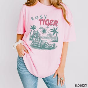 May include: Pink t-shirt featuring a graphic of a tiger wearing sunglasses and relaxing on a beach chair. The text "Easy Tiger" is printed on the shirt.