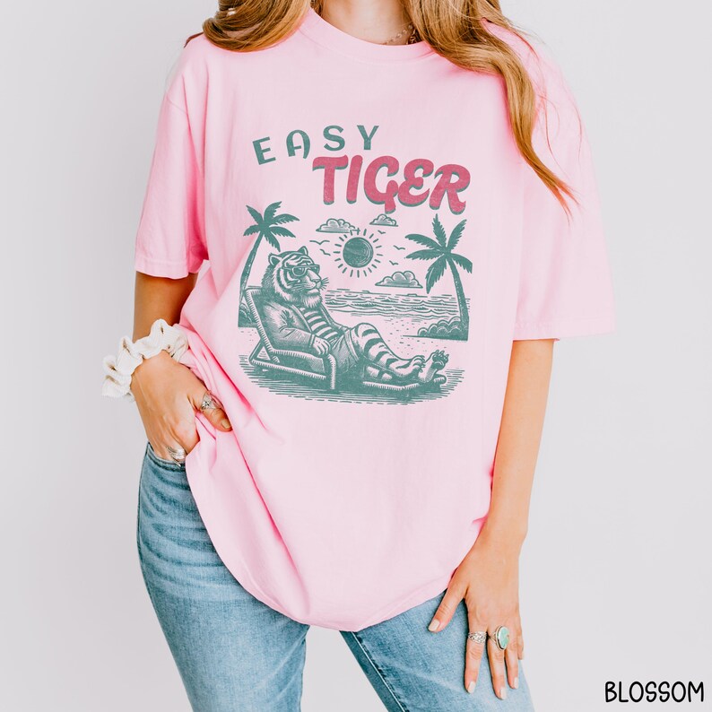 May include: Pink t-shirt featuring a graphic of a tiger wearing sunglasses and relaxing on a beach chair. The text "Easy Tiger" is printed on the shirt.