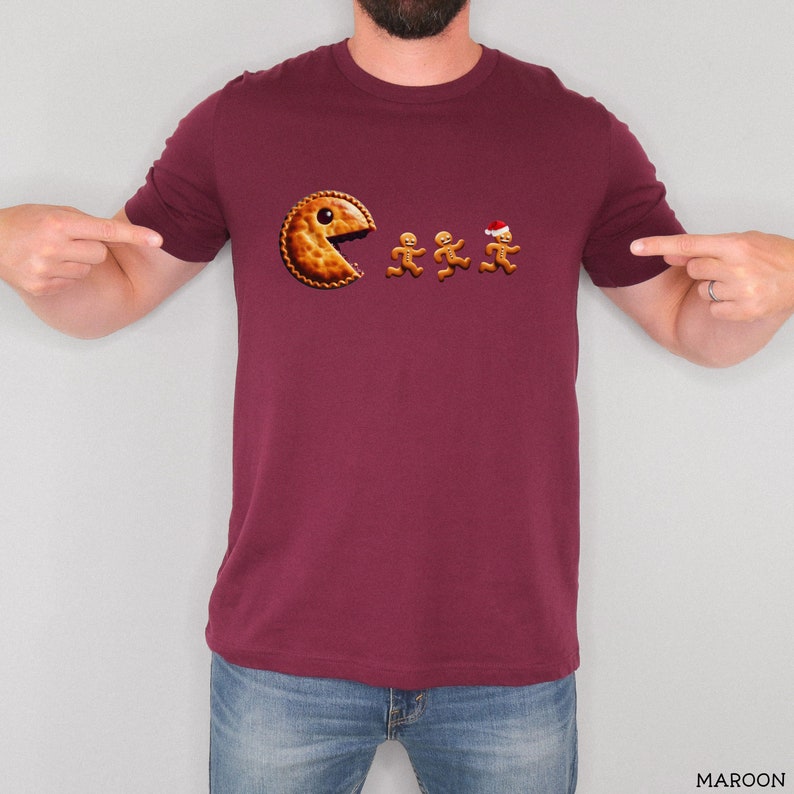 Pu&ograve; includere: Una t-shirt a maniche corte color bordeaux con una stampa grafica che raffigura una torta dorata a forma di personaggio di un videogioco che insegue tre biscotti di pan di zenzero, uno dei quali indossa un piccolo cappello da Babbo Natale.