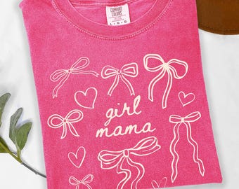 May include: A hot pink short-sleeve t-shirt with a crew neckline and a Comfort Colors brand label. The front features a white graphic print of multiple bows and hearts with the text "girl mama" written in a cursive script in the center.