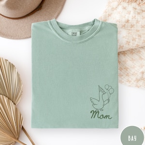 Op de afbeelding: Een lichtgroen Comfort Colors T-shirt met een cartoon gans met een feesthoedje en ballonnen. Het woord "Mom" staat onder de gans in een scriptlettertype. Het shirt is netjes opgevouwen.