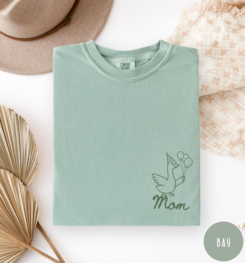 Op de afbeelding: Een lichtgroen Comfort Colors T-shirt met een cartoon gans met een feesthoedje en ballonnen. Het woord "Mom" staat onder de gans in een scriptlettertype. Het shirt is netjes opgevouwen.