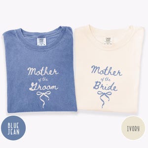 May include: Two t-shirts, one blue jean and one ivory, with the words "Mother of the Groom" and "Mother of the Bride" in script. Each shirt has a bow graphic below the text. The shirts are folded.