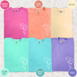 May include: Six folded t-shirts in various colors: pink, orange, yellow, teal, turquoise, and purple. Each shirt has a white balloon graphic with initials. Colors are labeled: Crunch-Berry, Terra-Cotta, Butter, Island Reef, Chalky Mint, and Violet.
