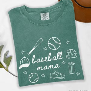Puede incluir: Camiseta verde claro con un gr&aacute;fico blanco de tem&aacute;tica de b&eacute;isbol que dice "baseball mama". El gr&aacute;fico incluye una pelota de b&eacute;isbol, un bate, un guante, tacos, palomitas de ma&iacute;z y una gorra de b&eacute;isbol.