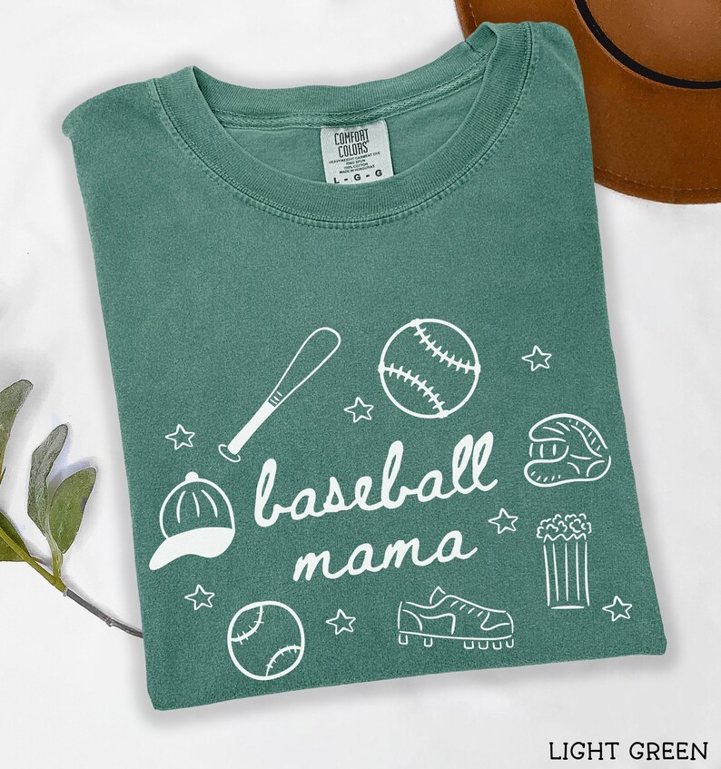 Puede incluir: Camiseta verde claro con un gr&aacute;fico blanco de tem&aacute;tica de b&eacute;isbol que dice "baseball mama". El gr&aacute;fico incluye una pelota de b&eacute;isbol, un bate, un guante, tacos, palomitas de ma&iacute;z y una gorra de b&eacute;isbol.
