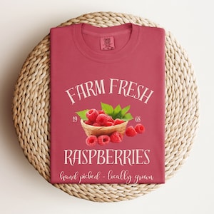 May include: A red t-shirt with the text "FARM FRESH RASPBERRIES hand picked - locally grown" in white. A basket of red raspberries is pictured in the center of the shirt.