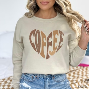May include: A sand-colored sweatshirt featuring a heart-shaped design with the word "COFFEE" in brown and tan tones. The sweatshirt has a crew neck and long sleeves, perfect for casual wear. The word "SAND" is printed at the bottom right.