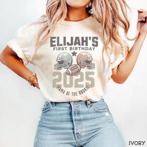 Puede incluir: Una camiseta de manga corta color marfil con un gr&aacute;fico que incluye el texto ELIJAH'S FIRST BIRTHDAY 2025 MAMA OF THE ROOKIE, dos cascos de f&uacute;tbol americano y un bal&oacute;n. El dise&ntilde;o est&aacute; centrado en el pecho con un estilo vintage desgastado.