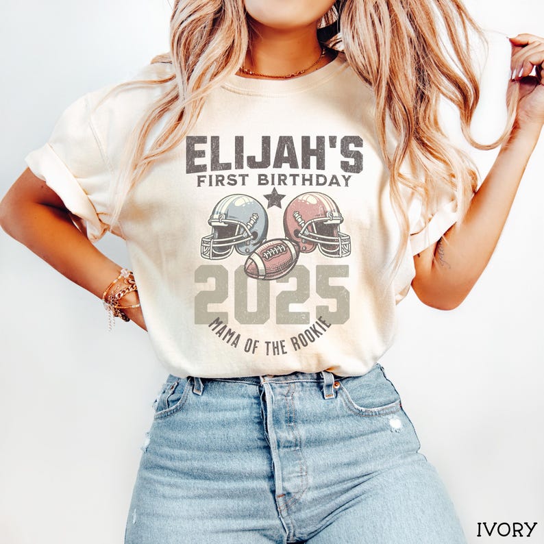 Puede incluir: Una camiseta de manga corta color marfil con un gr&aacute;fico que incluye el texto ELIJAH'S FIRST BIRTHDAY 2025 MAMA OF THE ROOKIE, dos cascos de f&uacute;tbol americano y un bal&oacute;n. El dise&ntilde;o est&aacute; centrado en el pecho con un estilo vintage desgastado.