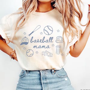 Puede incluir: Una camiseta blanca con un dise&ntilde;o gr&aacute;fico azul que dice "baseball mama" e incluye ilustraciones con tem&aacute;tica de b&eacute;isbol como una pelota de b&eacute;isbol, un bate, un guante, un sombrero y tacos. La camiseta la lleva puesta una persona con el pelo largo y rubio y va combinada con unos vaqueros azules.