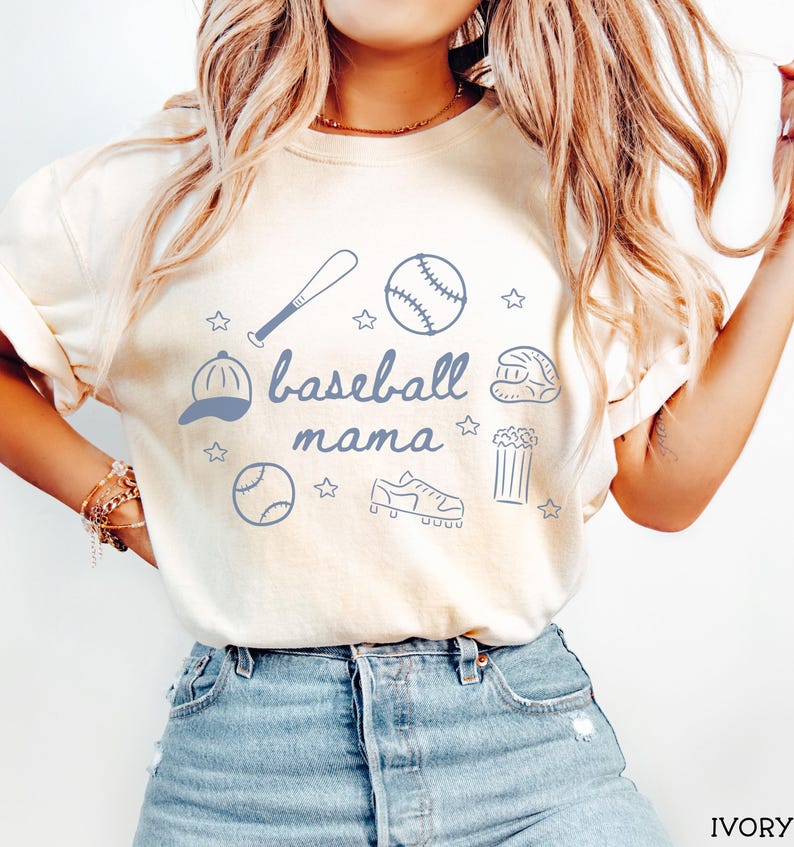 Puede incluir: Una camiseta blanca con un dise&ntilde;o gr&aacute;fico azul que dice "baseball mama" e incluye ilustraciones con tem&aacute;tica de b&eacute;isbol como una pelota de b&eacute;isbol, un bate, un guante, un sombrero y tacos. La camiseta la lleva puesta una persona con el pelo largo y rubio y va combinada con unos vaqueros azules.