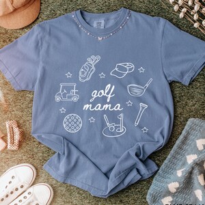 Puede incluir: Una camiseta azul vaquero con un dise&ntilde;o gr&aacute;fico blanco de equipo de golf y el texto "golf mama".