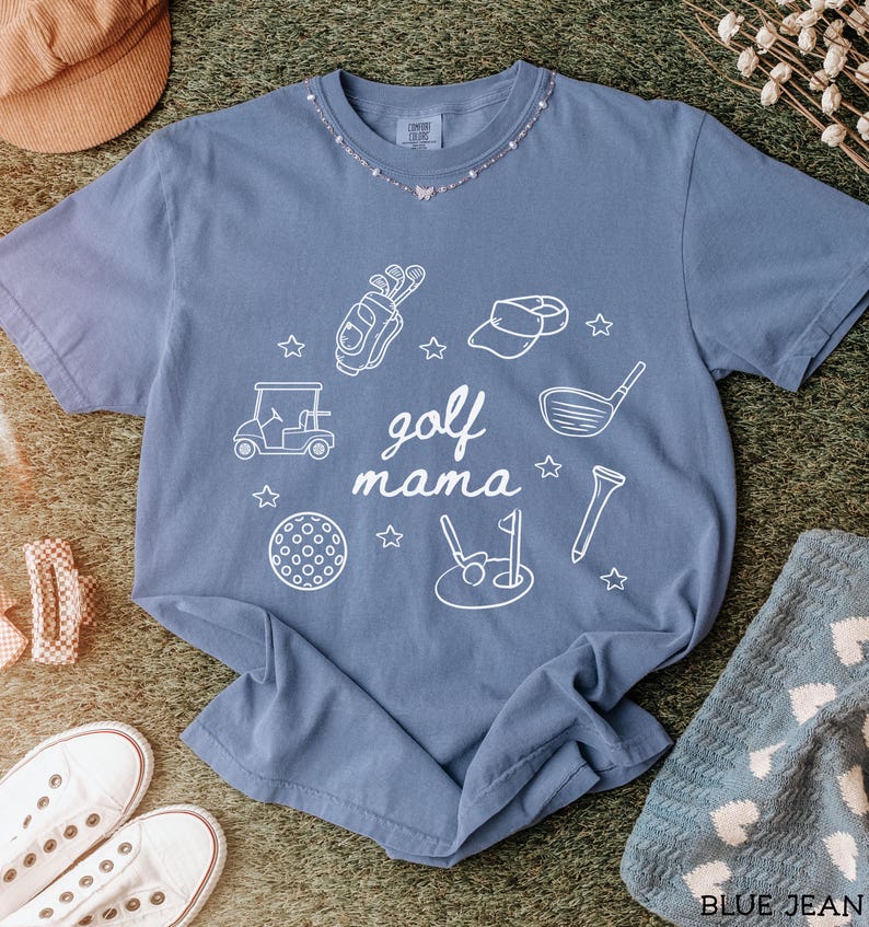 Puede incluir: Una camiseta azul vaquero con un dise&ntilde;o gr&aacute;fico blanco de equipo de golf y el texto "golf mama".