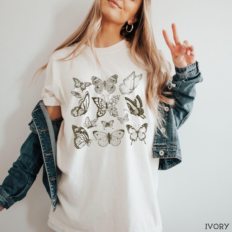 May include: A white t-shirt with a pattern of black butterflies. The butterflies are arranged in a grid pattern and are all different sizes and shapes. The shirt is a relaxed fit and has a crew neck.