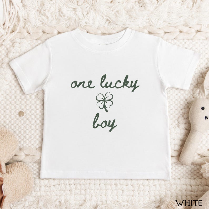 May include: White toddler t-shirt with the text "one lucky boy" in dark green script, featuring a four-leaf clover graphic. The shirt is short-sleeved and made of a soft material.