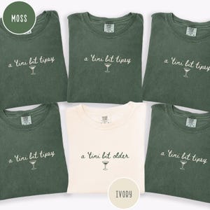 May include: A collection of moss green and ivory t-shirts. The shirts feature the phrase "a 'tini bit tipsy" and a martini glass graphic, with one shirt displaying "a 'tini bit older". The shirts are folded and displayed together.