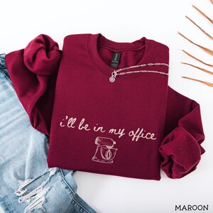 May include: A maroon long-sleeve crewneck sweatshirt featuring the white text "I'll be in my office" written in a cursive script above a line-art illustration of a stand mixer. The sweatshirt is paired with distressed blue denim jeans.