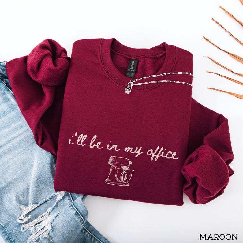 May include: A maroon long-sleeve crewneck sweatshirt featuring the white text "I'll be in my office" written in a cursive script above a line-art illustration of a stand mixer. The sweatshirt is paired with distressed blue denim jeans.