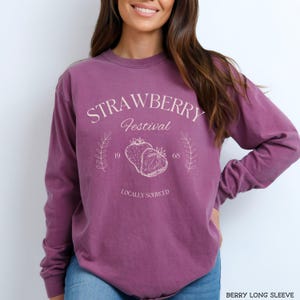 K&ouml;nnte beinhalten: Ein lilafarbenes Langarmshirt mit dem Text "Strawberry Festival" und einer Grafik von zwei Erdbeeren. Der Text "Locally Sourced" befindet sich unter der Grafik.