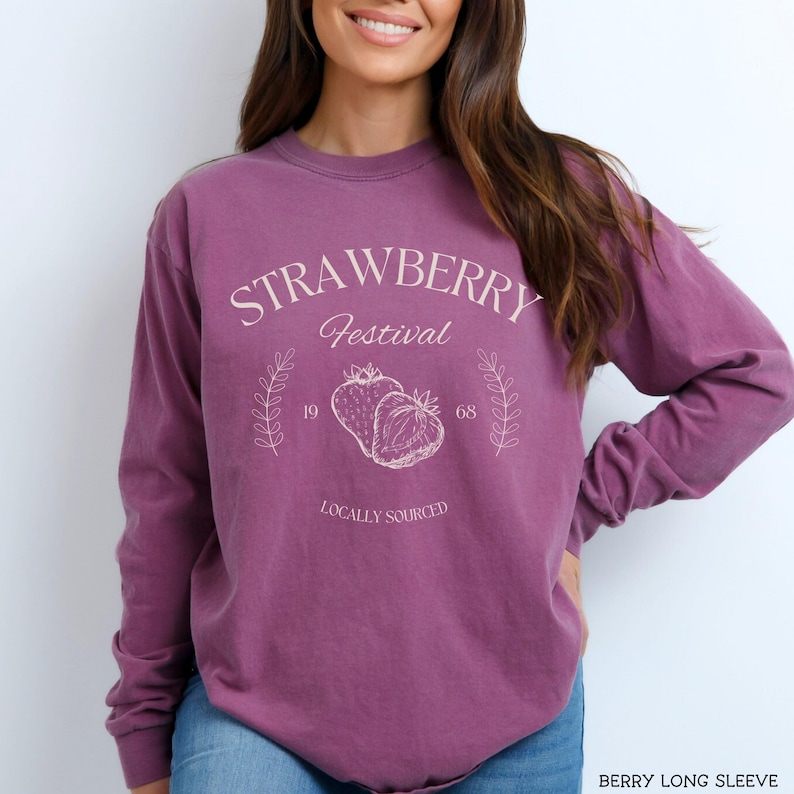 K&ouml;nnte beinhalten: Ein lilafarbenes Langarmshirt mit dem Text "Strawberry Festival" und einer Grafik von zwei Erdbeeren. Der Text "Locally Sourced" befindet sich unter der Grafik.