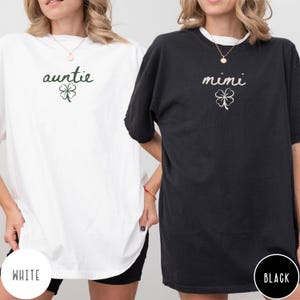 May include: Two oversized t-shirts, one white and one black. The white shirt has the word "auntie" and a shamrock graphic. The black shirt has the word "mimi" and a shamrock graphic. The word "WHITE" is printed on the bottom left of the white shirt, and "BLACK" is printed on the bottom right of the black shirt.