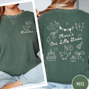Op de afbeelding: Mosgroen sweatshirt met witte afbeelding van een gans met ballonnen en de tekst "Grandma" op de voorkant. De achterkant heeft een gans met een feesthoed, taart en de tekst "Olivia's One Silly Goose".