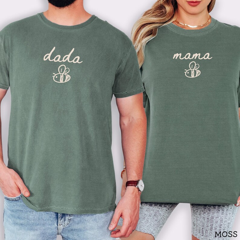 May include: Two moss green t-shirts. One shirt has the word "dada" with a bee graphic, the other says "mama" with a bee graphic. The shirts are short-sleeved and made of a soft material.