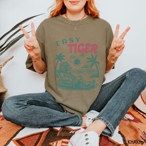 May include: A khaki green t-shirt featuring a graphic of a tiger relaxing on a beach with palm trees and the text "EASY TIGER" in pink.