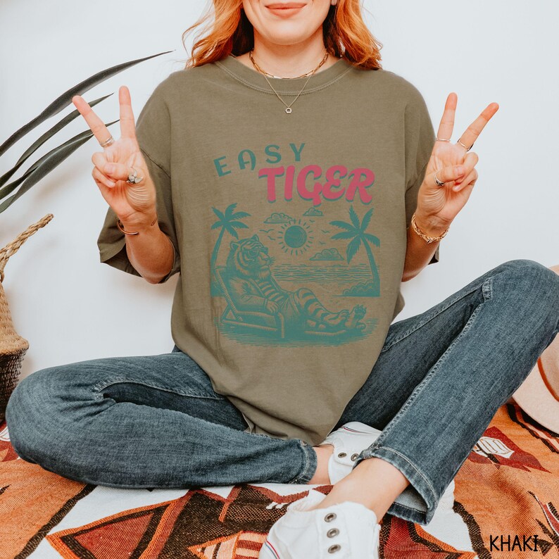 May include: A khaki green t-shirt featuring a graphic of a tiger relaxing on a beach with palm trees and the text "EASY TIGER" in pink.