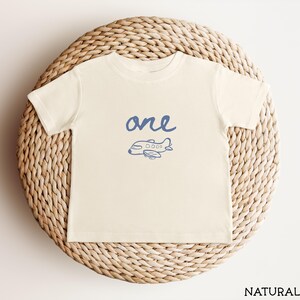 以下が含まれることがあります： クリーム色の半袖Tシャツ。胸の中央に小さな飛行機の青いグラフィックプリントと、手書き風の「one」という文字がデザインされています。クルーネックで、裾はシンプルなストレートカットのシルエットです。