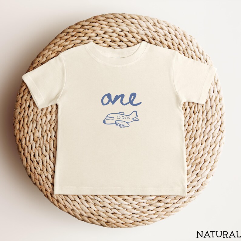 以下が含まれることがあります： クリーム色の半袖Tシャツ。胸の中央に小さな飛行機の青いグラフィックプリントと、手書き風の「one」という文字がデザインされています。クルーネックで、裾はシンプルなストレートカットのシルエットです。