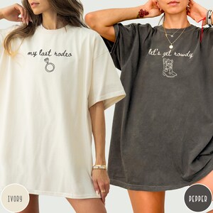 May include: Two short-sleeve t-shirts in ivory and pepper gray. The ivory shirt features the text "my last rodeo" with a diamond ring graphic. The pepper gray shirt features the text "let's get rowdy" with a cowboy boot graphic in white ink.